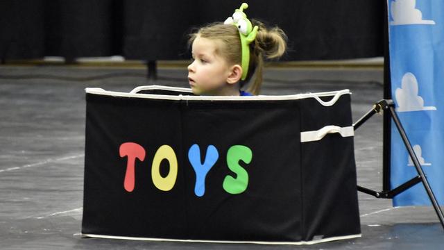 A young child with a green 3-eyed alien Toy Story headband sits inside a black box labeled "TOYS."