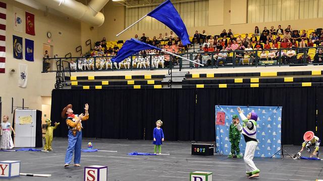 A performance scene featuring several characters in costumes, including Buzz Lightyear, Woody, and other Toy Story characters, interacting on a stage. Two performers are tossing large blue flags, while an audience watches from the stands.