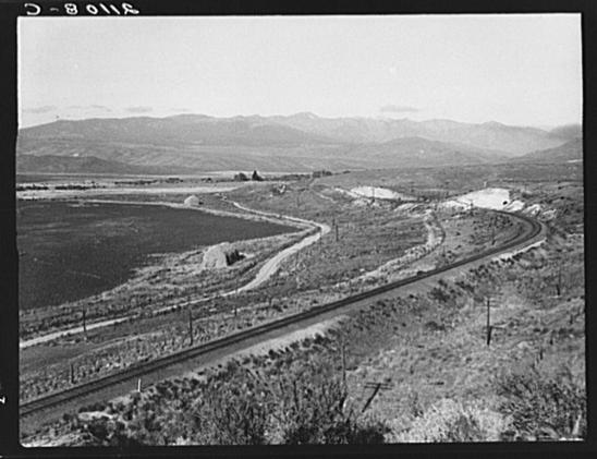 The image is a black and white photograph depicting an expansive rural landscape. In the foreground, there's a railway track that cuts through the middle of the frame from left to right, with sparse vegetation on both sides. The terrain appears dry and arid in certain areas, while other patches look slightly more cultivated or maintained.
In the midground, we see winding roads intersecting each other, leading towards distant hills and mountains under a clear sky. There's an open field that extends into the background where some buildings are visible at a distance on elevated ground. The overall atmosphere is one of desolation with no immediate signs of human activity or presence.
The landscape suggests a rural setting in Eastern Oregon as indicated by the accompanying text, which also specifies it to be Small Finger Valley along U.S. 30, Baker County, Oregon. A date stamp from 'D-8' on top left and reference code '6495b830ba25398d90c500d2.jpg' are visible in the image metadata.
This photograph appears to be part of a larger collection documenting agricultural practices or conditions, as hinted by another caption stating "Farmers have just sown their winter wheat."