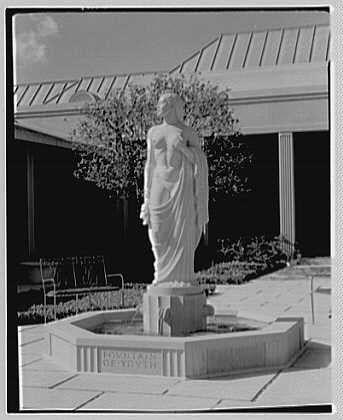 A black and white photo shows a statue of an elegant woman, draped in flowing clothing. She stands on a stone pedestal with engraved lettering around its base. The background features manicured landscaping, including shrubs and trees, as well as the architectural elements like tiled flooring and pillars typical of formal gardens or courtyards. This serene scene likely captures the ambiance of a sophisticated outdoor space, possibly part of an estate or public institution such as a museum or gallery.