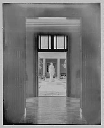 The image depicts a monochrome photograph of an architectural interior, likely taken during the early 20th century. The perspective is from within a hallway or corridor that leads to another room through double doors.
Through these open doors, we see what appears to be a statue or sculpture positioned in front of a set of windows with horizontal panes, allowing natural light into the space. On either side of this central area are tall pillars or columns which support the structure above and add symmetry to the composition.
The scene is empty aside from the distant figure near the windowed door, giving it an air of solitude. The photograph's black-and-white nature emphasizes contrasts between shades of grey, enhancing textures like that of walls, floor tiles, and possibly a tiled patio or courtyard area beyond the doors visible through reflections on glass surfaces.