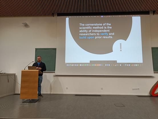 Ben Hermann talking from a podium, with a slide in the background that reads "The cornerstone of the scientific method is the ability of independent researchers to verify and build upon prior results."