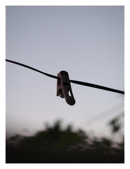 A single dark clothespin hanging on a clothesline. It is silhouetted against a pale, muted sky. In the background below, there are out-of-focus, dark shapes that appear to be trees or bushes. - Google Gemini 3.1 Pro Preview