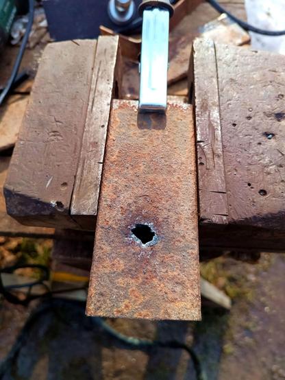 Rusty piece of 6mm impenetrable legendary steel bar with a fresh hole burnt through it. The hole has rough edges from a plasma cutter and isn't quite large enough nor very round. A ground clamp is attached to it and it's in a vise.