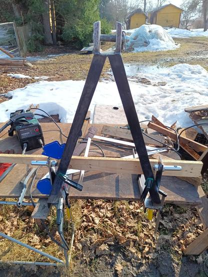 The pallet forks being worked on, clamped to the improvised outdoor workbench so they don't move. The top fitting is welded on, two C-channel pieces with angled tops and the sides shortened. A pin is going through them. Further down two tangs with link arm pins in them are tacked on, one still being held by blue welding magnets. A piece of wood with pencil markings is clamped to the steel triangle as a guide. A small green welder stands on the bench behind. The yard is one giant puddle of mud and melting snow.