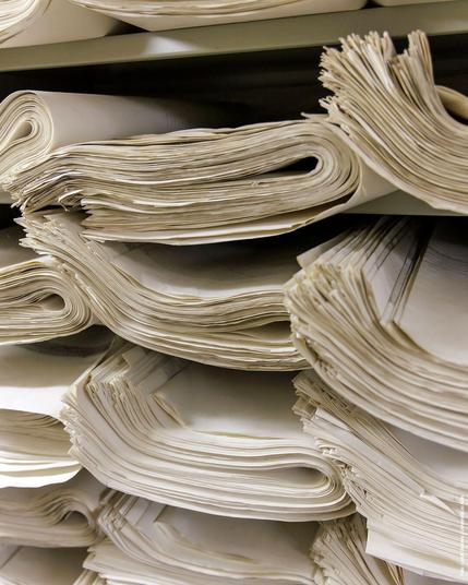 Gestapelte, aufgerollte großformatige Papierbögen in einem Archivregal.