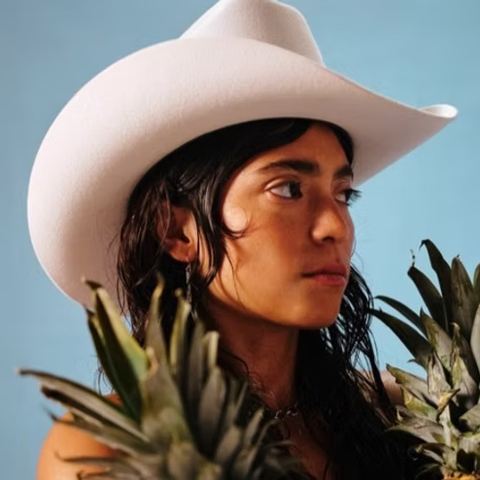 A Latina woman with straight, dark hair looks to the right. She stands against a pale blue sky and wears a white cowboy hat. There are two pineapple stems in the foreground.