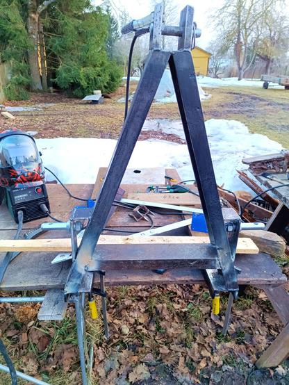 The same beam, now welded in between the triangular supports of the homemade palletforks. The ears are now also complete, with flat bars forming an angle to support the connection. A welding helmet is on the workbench and a clamp attached at the top.