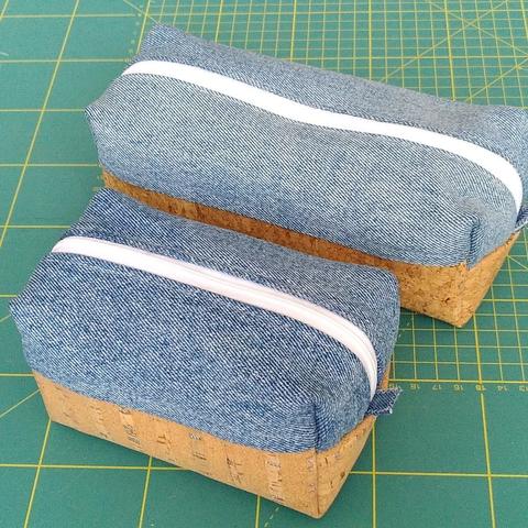 Deux trousses rectangulaires en volume. L'une est plus courte que l'autre. Elles sont posées sur un tapis de coupe de couturière. Faites en deux tissus différents. La partie supérieure est en jeans recyclé et la partie inférieure en liège. La fermeture éclair est blanche.