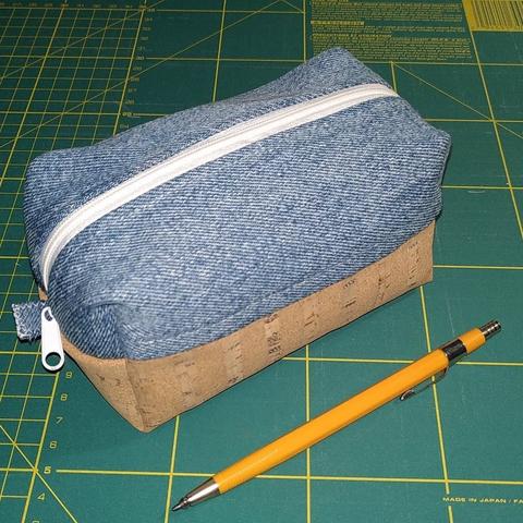 Trousse rectangulaire en volume. Elle est posée sur un tapis de coupe de couturière. Faite en deux tissus différents. La partie supérieure est en jeans recyclé et la partie inférieure en liège. La fermeture éclair est blanche. Un crayon porte-mine est posé à côté pour se rendre compte de la dimension.