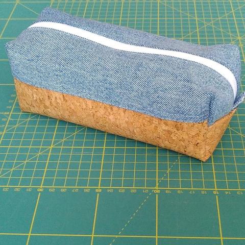 Trousse rectangulaire en volume. Elle est posée sur un tapis de coupe de couturière. Faite en deux tissus différents. La partie supérieure est en jeans recyclé et la partie inférieure en liège. La fermeture éclair est blanche.