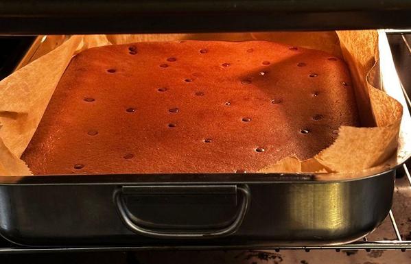 A large rectangular banana bread with chocolate chips baking in an oven inside a metal roasting pan lined with parchment paper. The top is evenly browned and dotted with small chocolate chips across the surface.