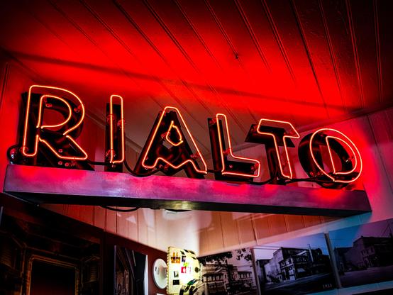A close-up, low-angle shot of a red neon sign that spells out "RIALTO" in all caps. The sign is mounted on a gray metal base. The glowing red light from the neon tubes reflects onto the white wooden-slat ceiling above. In the background below the sign, several framed black-and-white historical photographs of a theater and a city street from 1933 are partially visible. The overall mood is warm, retro, and cinematic.