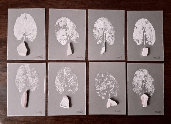 8 PostkARTenUnikate auf grausilberfarbenem Grund mit silberfarbener Umrandung, jeweils mit einem weißem Abdruck eine Meermangold-Blattes, das aus einem Scherbenfundstück "wächst".
Tiny Art by Teena