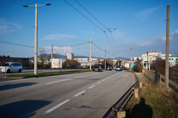 Eine Bushaltestelle an einer 2 Spurigen Straße. Schmaler Gehsteig, es geht eine Rampe zur Seite runter. Es schaut verwachsen und alt aus.
