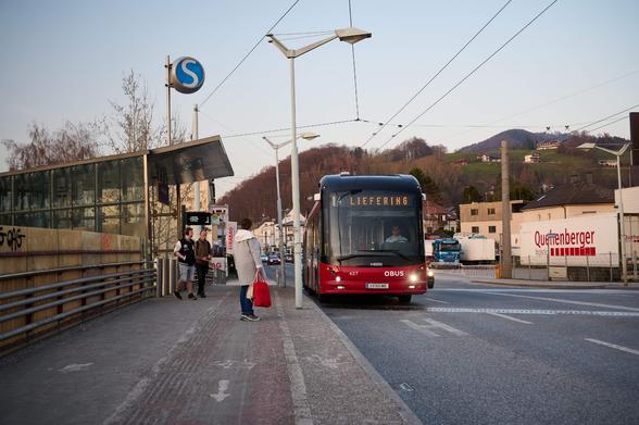 Ein Bus steht neben einem breiten Gehsteig mit Blindenleitsystem. Es warten Leute auf den Bus. Es ist ein Radweg zu erkennen. Links ist ein Eingang zu einer S-Bahn Station zu erkennen.