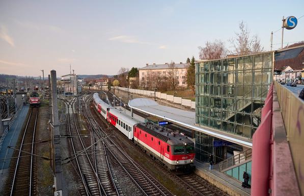 Blick von der Brücke runter auf einen Zug. Man erkennt das Stiegenhaus der S-Bahn Station von der Brücke runter. Die S-Bahn Station schaut recht modern aus und ist mit viel Glas gebaut.