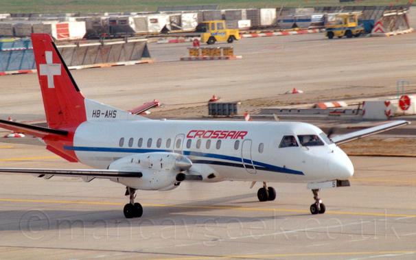 Side view of a twin propellor-engined airliner taxiing from left to right.
The plane is largely white, with a thin blue and black stripe running along the body, and red "CrossAir" titles on the upper forward fuselage.
A diagonal red band wraps around the rear fuselage and tail, with a large white cross on the upper half of the tail, and the black registration "HB-AHS" on a white fillet at the front of the tail, above the rear fuselage.
Grey concrete apron fills the foreground and most of the background, with rows of luggage containers and a pair of yellow baggage tugs at the top of the frame.