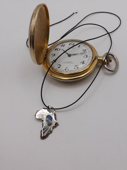 a silver pendant in shape of Africa! it has a light blue small gemstone in the middle and black cord. an old pocket watch in the background