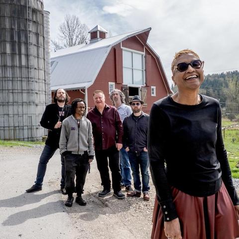 In the foreground, an older Black woman with short red hair smiles into the camera. She wears tinted glasses, a black shirt, and red skirt. In the background, a group of five men laugh into the camera. Behind them is a red barn and green field.