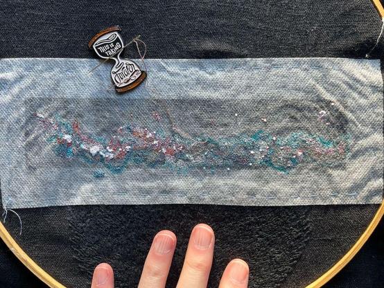 Lia’s fingers show a wooden embroidery hoop with black linen with a rectangle of dissolvable material on which is printed the Milky Way in black and white with some red and blue markings. Some tiny single strand seed stitch and French knots in white, peach, oranges, aubergine, and dark browns and grays have been stitched. The tiny needle holds dusky rose thread. Below the Milky Way is black on black whipped backstitch that looks a bit like flames, it’s pubic hair. An hourglass-shaped needle minder that reads “this is taking forever” is attached to the cloth.