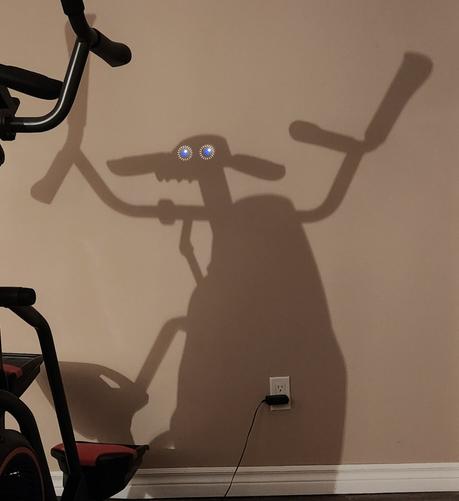 Shadow of an exercise machine on a beige wall. It kind of looks like a nerdy alien sheep. Eyes have been digitally added to make it more obvious.