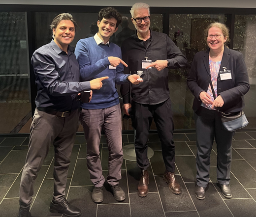 The FIZ Karlsruhe KDAI winner team with (from left to right): Hossein Beygi Nasrabadi, Ebrahim Norouzi, Harald Sack, Heile Fliegl, standing in the Foyer of the meeting venue in Siegburg. Ebrahim is holding a miniature trophee in his left hand. Eberybody is pointing with their index fingers on the trophee.