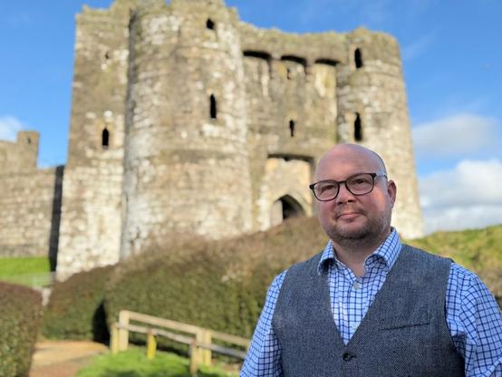 A bald man wearing glasses, a blue checked shirt and a grey herringbone waistcoat stands in front of the stone keep of a ruined medieval castle on a bright, sunny day.