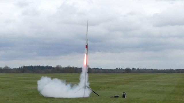 Ein Minisatellit startet auf einer grünen Wiese, man sieht Feuer und Qualm. Im entfernten Hintergrund sind Bäume zu sehen.