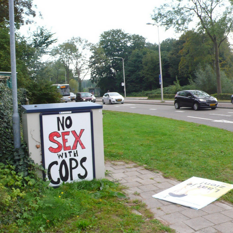 straat kastje langs auto weg met gras en bomen, rijdende auto's en een bus. Het vorig reclame bord ligt op de grond
Zwart met rode letters