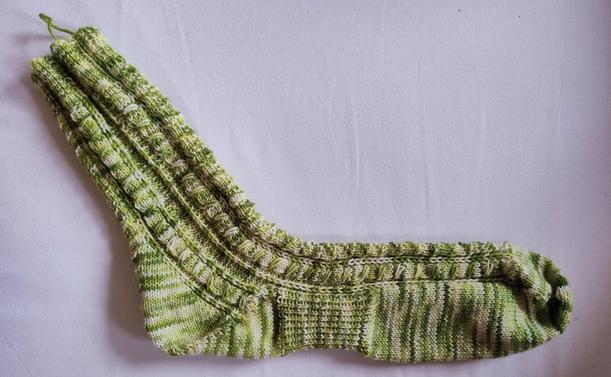 A hand-knit sock on a white background.
The sock is made from green white variegated yarn. Pattern is textured with cables.
