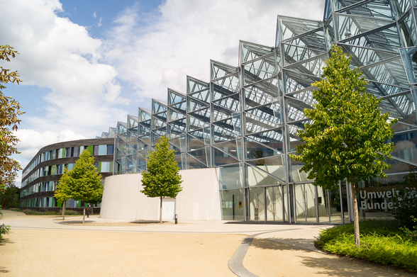 Außenansicht des Umweltbundesamtes in Dessau: Modernes Gebäude mit großer Glas-Stahl-Konstruktion und markanter, dreieckiger Dachstruktur. Vor dem Eingang stehen mehrere Bäume entlang eines hellen Weges; im Hintergrund ist ein teilweise bewölkter Himmel zu sehen.