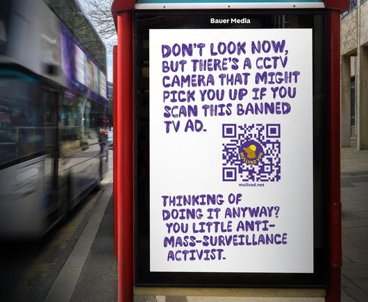 An image of the advert on the side of a bus stop.
The background is white, with purple stylised text which reads: "Don't look now, but there's a CCTV camera that might pick you up if you scan this banned TV ad."
Below is a QR code which leads to mullvad.net, the QR code is purple, and has the Mullvad VPN logo and mole inside.
Underneath is more text which reads: "Thinking of doing it anyway? You little anti-mass-surveillance activist."
