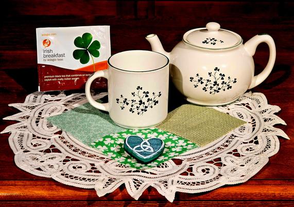 White and green tea tableau on Irish Lace with a quilting square of green prints including shamrocks. The teapot and mug are white rimmed in green and with a dainty spray of shamrocks at center. There is a wooden heart painted green with a white Celtic triskele design handpainted on it. The tea is Irish Breakfast black loose-leaf tea from Adagio.
