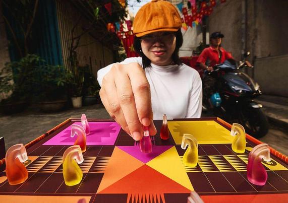 A hand moves a colorful game piece on a vibrant board, set in a lively street decorated with flags and a motorcycle in the background.