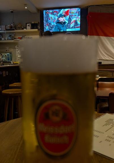 Foto des gemütlichen Innenraums einer Gaststätte mit Holztisch und Barhockern. Im Vordergrund steht ein großes Bierglas mit weißem Schaum und rotem Etikett „Reissdorf“ auf einem Tisch. Im Hintergrund läuft auf einem Fernseher das Fußballspiel VfB Stuttgart - FC Porto, im Bild VfB-Trainer Sebastian Hoeneß vor den Fans der Cannstatter Kurve. Daneben eine rot-weiße Flagge an der Wand und Regale mit Gläsern.