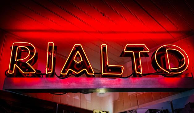 A close-up, low-angle photograph of the word "RIALTO" in red neon letters. The glowing glass tubes follow the contours of dark, three-dimensional block letters. The vibrant red light illuminates the white-painted wooden ceiling in the background, creating a soft glow that contrasts with the sharp lines of the neon. The perspective makes the letters appear large and imposing, capturing the industrial and artistic details of the vintage sign.