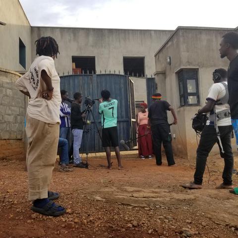 A small film crew records a scene outside a studio compound, with a camera on a tripod and a boom microphone pointed toward an actor standing near a metal gate, while other crew members watch and assist with equipment.
