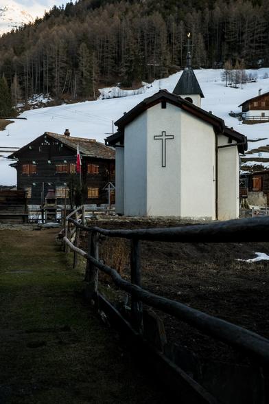 Auf einem Wanderweg: Im Hintergrund ein noch eingeschneiter Hang, bewaldet mit Lärchen und Tannen.Bildmitte ein typisches Walliser Holzchalet mit roten Fensterläden und Steinplattendach, direkt daneben eine Kapelle mit dem Kreuz gut sichtbar. Man sieht noch einen Holzzaun, der direkt am Wegesrand steht - das Foto ist so aufgenommen, dass es wirkt, als blicke man direkt vom Wanderweg in die Szenerie.