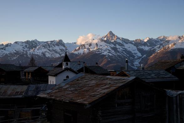 Blick über einen alten Walliser Weiler - über die Steinplattendächer hinweg, direkt auf das Panorama mit Bietschhorn. Das Bietschhorn ist ein markanter Berg; sehr typisch ist seine pyramidenförmige Ausprägung. In der Bildmitte ist eine Kapelle zu sehen und die ganze Szene präsentiert sich im letzten Sonnenlicht; die Häuser sind bereits im Dunkel des Schattens untergetaucht.