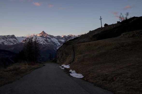 Blick auf das Bietschorn im Abendlicht. Der Blick führt vom unteren Bildrand einer Strasse bergab, entlang in Richtung des Bietschhornpanoramas. Auf der rechten Bildseite ist ein Bergrücken - darauf steht ein Kreuz aus Holz im Schatten und überragt die Szenerie bewachend. Die Bergkette um das Bietschorn ist noch ziemlich eingeschneit und auch am Strässchen, von wo aus das Bild gemacht wurde, hat es letzte Schneereste.