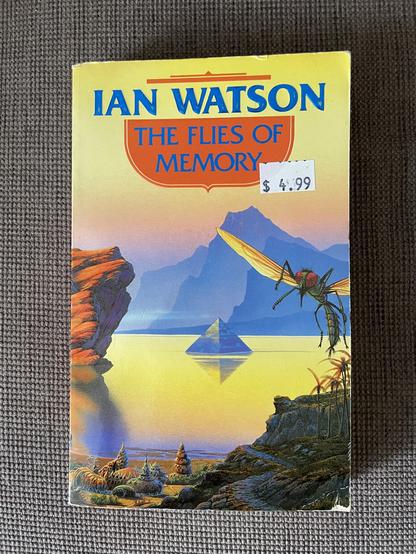 A photo of a paperback edition of the novel The Flies of Memory by Ian Watson which features an otherworldy landscape, predominantly yellow, with a giant flying insect in the foreground.