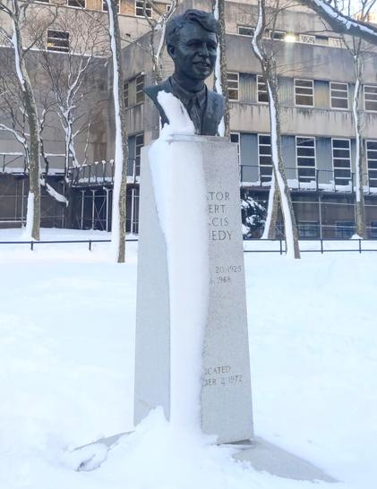 Robert F. Kennedy memorial bust at Brooklyn Borough Hall Park. The ground and left side of the culumn are covered in snow. The snow goes up to the left side of the Kennedy bust's shoulder, but nowhere else.