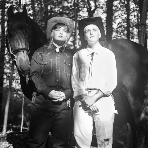 A and white photo of two people standing in a forest with a horse. A beam of sunlight hits them directly. The person on the left is a white person in a white cowboy hat, shoulder-length bleach blond hair braided into plaits, and a dark denim jacket. The person's hands are clasped. On the right is a blonde person in a black cowboy hat, light denim shirt, and light denim pants. They are holding their wrist with one hand in front of them.