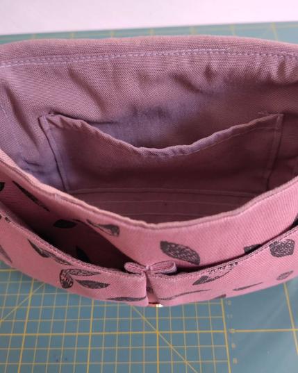 Petit sac à main avec rabat. Il est ouvert et on voit qu'à l'intérieur il y a une poche, deux autres l'une à côté de l'autre sont cousues à l'extérieur. Le tissu est rose avec des feuilles noires comme imprimées au tampon. Il a une sangle. Le sac est posé sur mon tapis de coupe classique (il est vert avec des lignes jaunes qui marquent les centimètres)