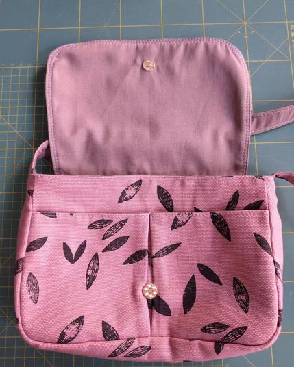 Petit sac à main avec rabat qui est ouvert et l'on voit deux poches l'une à côté de l'autre cousues à l'extérieur sur la poche principale ainsi que le bouton en métal aimanté, il est couleur cuivre. Le tissu est rose avec des feuilles noires comme imprimées au tampon. Il a une sangle. Le sac est posé sur mon tapis de coupe classique (il est vert avec des lignes jaunes qui marquent les centimètres)