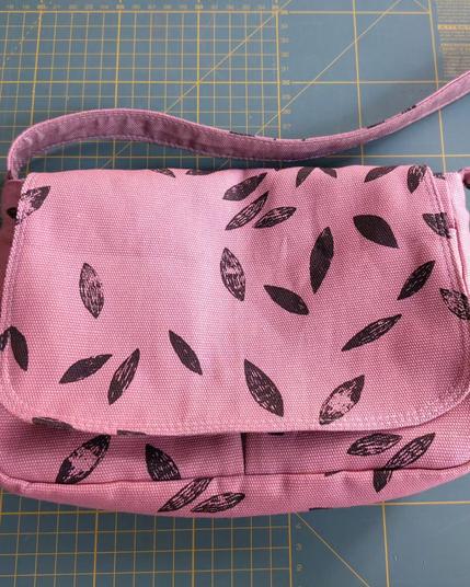 Petit sac à main avec rabat. Le tissu est rose avec des feuilles noires comme imprimées au tampon. Il a une sangle. Le sac est posé sur mon tapis de coupe classique (il est vert avec des lignes jaunes qui marquent les centimètres)