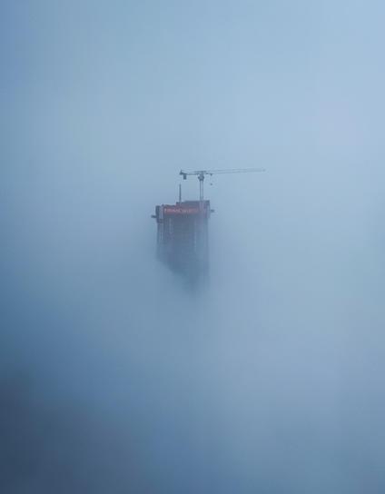 The very top of a sky scraper under construction with a crane on top. It is poking out of a heavy cloud bank.