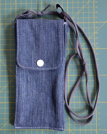 Sacoche pour téléphone mobile faite avec des restes de jeans avec un lacet de chaussure gris en guise de sangle et et un rabat avec un bouton pression blanc. Elle est posée sur mon tapis de coupe classique (il est vert et on voit les lignes jaunes qui indiquent chaque centimètre)
