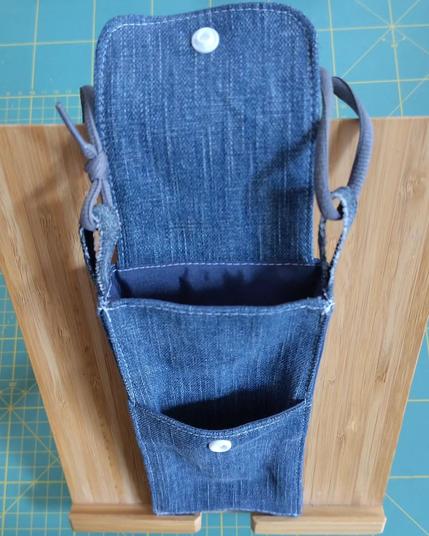 Sacoche pour téléphone mobile faite avec des restes de jeans avec un lacet de chaussure gris en guise de sangle et un rabat avec un bouton pression blanc. Elle est ouverte, on voit qu'elle a une poche extérieure. Elle est posée sur un support en bois de bambou qui est sur mon tapis de coupe classique (il est vert et on voit les lignes jaunes qui indiquent chaque centimètre)